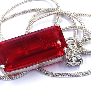 May include: A rectangular, faceted red gemstone pendant set in a silver-toned metal frame with a decorative bail, attached to a thin, flexible silver-toned snake chain necklace that loops around the pendant in a circular arrangement.
