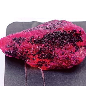 May include: A large, rough, pink ruby gemstone with black inclusions. The ruby is on a dark gray surface.