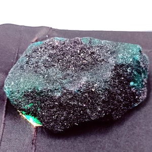 May include: A rough, dark green emerald gemstone with a slightly irregular shape. The stone has a textured surface and is set against a black background.