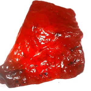 May include: A raw, irregularly shaped piece of vibrant red mineral with a glossy, translucent surface. The stone features deep crimson hues, natural facets, and small dark inclusions scattered throughout its crystalline, textured structure.