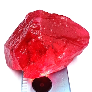 Puede incluir: Una gema grande, en bruto, de color rojo rubí con una superficie texturizada. La piedra tiene forma irregular y parece en bruto y sin pulir. La gema está sobre un fondo blanco.