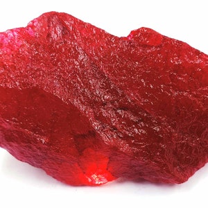 May include: A large, rough, red ruby gemstone. The ruby has a deep, rich red color and a slightly translucent appearance.