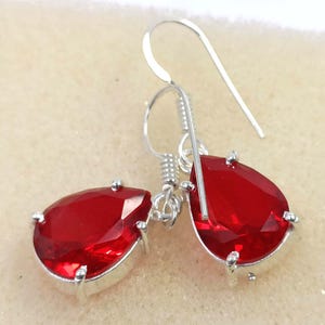 May include: A pair of silver drop earrings featuring large, teardrop-shaped, faceted red gemstones. The gemstones are set in a silver bezel and suspended from a silver hook. The earrings are displayed against a neutral background.