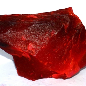May include: A rough-cut, deep red gemstone with a textured surface. The stone has a rich, saturated color, with variations in tone creating depth. The overall shape is irregular, showcasing its natural, unpolished state. The gemstone is likely a ruby or garnet.