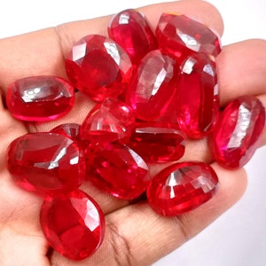May include: A collection of oval-shaped, faceted, red gemstones. The gemstones are arranged in a pile and appear to be loose.