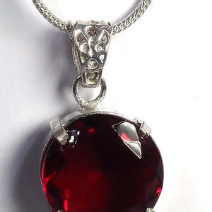 May include: A silver pendant necklace featuring a large, round, deep red gemstone. The gemstone is held in place by four silver prongs. The pendant hangs from a delicate silver chain. The setting and chain are silver.