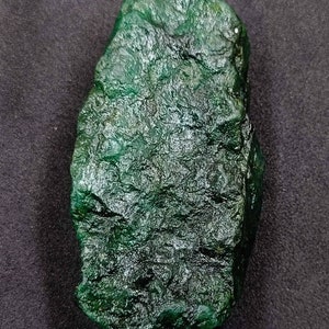 May include: A rough-cut, deep green emerald gemstone. The surface has a textured, uneven appearance with variations in color and tone. The emerald is displayed against a dark background.