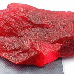 May include: A large, rough, red ruby gemstone. The ruby is a deep red color and has a natural, unpolished finish.