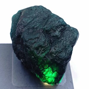 May include: A rough, uncut emerald gemstone with a deep green color. The surface has a textured appearance, with areas of dark green and bright, glowing green. The gemstone is displayed on a light gray surface.