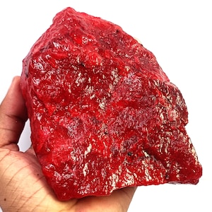 Könnte beinhalten: Ein großes, rohes und unregelmäßig geformtes Mineralexemplar in einer leuchtend tiefroten Farbe. Die Oberfläche des Steins weist eine glänzende, kristalline Textur mit natürlichen Graten, Spalten und Variationen in der Lichtreflexion auf.
