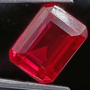 Puede incluir: Una piedra preciosa de color rojo intenso con un corte esmeralda rectangular. La piedra preciosa está sujeta por unas pinzas.