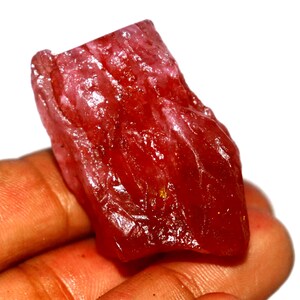 May include: A raw, unpolished gemstone with a deep red color and a translucent, crystalline texture. The stone features irregular, jagged edges and a natural, rough surface with visible internal fractures and light-reflecting facets throughout.