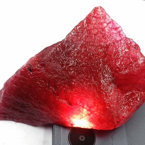 May include: A large, rough, red gemstone with a deep, rich color. The gemstone is illuminated from below, highlighting its internal structure.