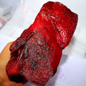 Könnte beinhalten: Ein großer, roher Rubin-Edelstein mit tiefroter Farbe und dunklen Einschlüssen. Der grob geschnittene Stein hat eine strukturierte Oberfläche und eine unregelmäßige Form, die seine natürliche Schönheit hervorhebt. Der Rubin wird in einer Hand gehalten.