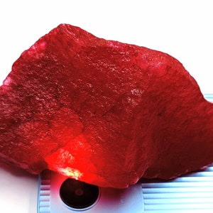 May include: A large, rough, red ruby gemstone. The ruby is illuminated from below, highlighting its deep red color.