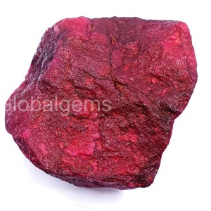 May include: A raw, uncut ruby gemstone in a deep red hue. The surface is textured and uneven, displaying visible crystalline formations. The text "globalgems" is visible on the left side of the stone.