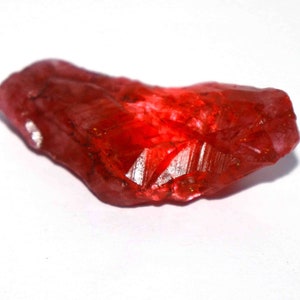 May include: A raw, unpolished gemstone with a vibrant, translucent red color. The stone features an irregular, elongated shape with sharp, angular facets and natural crystalline textures that catch the light, creating deep crimson highlights throughout.
