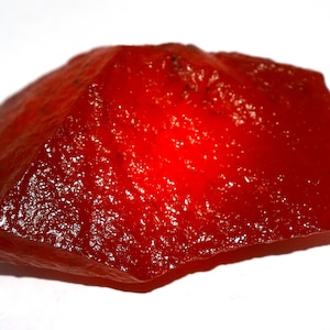 May include: A vibrant, translucent, red gemstone with a rough, uneven surface. The stone has a glossy appearance and is set against a white background. The shape is irregular, with a natural, organic form.