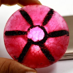 May include: A circular, translucent pink stone cabochon featuring a natural crackled texture throughout. The surface is marked with a dark black, six-pointed star pattern radiating from a central circle, creating a distinct contrast against the vibrant pink.