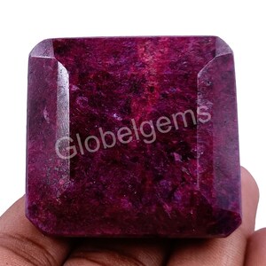 Op de afbeelding: Een dieprode edelsteen met een vierkante vorm en een facetgeslepen snit. De edelsteen heeft een ruwe, natuurlijke textuur en is gezet in een heldere, facetgeslepen zetting. De edelsteen is ongeveer 2,5 cm groot.