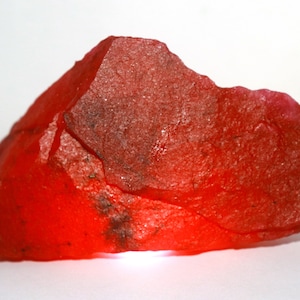 May include: A raw, translucent red mineral specimen with an irregular, jagged shape and a crystalline texture. The stone displays deep crimson hues with subtle variations in opacity and small dark inclusions visible across its rough surface.