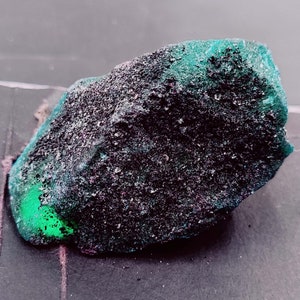 May include: A dark green and black rock with a rough, textured surface. The rock has a green hue with black speckles throughout.