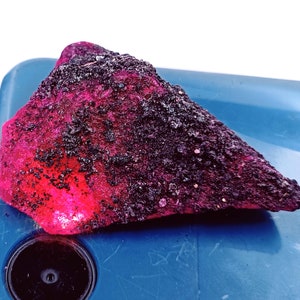 May include: A large, irregularly shaped, pink and black gemstone. The gemstone has a rough, textured surface and is likely a ruby.