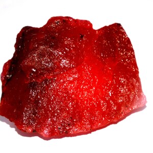 Op de afbeelding: Een ruw, doorschijnend rood mineraal exemplaar met een onregelmatige, grillige vorm. Het oppervlak vertoont een kristallijne, glasachtige textuur met diepe karmozijnrode tinten en kleine donkere insluitingen verspreid over de randen.