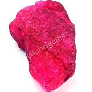 Op de afbeelding: Een grote, ruwe, roze robijn edelsteen. De robijn is een diepe, levendige roze kleur en heeft een natuurlijke, onbewerkte afwerking. De robijn is een ruwe, ongeslepen edelsteen.