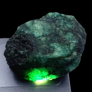 May include: A rough-cut emerald gemstone with a dark green hue and a textured surface. The bottom of the stone glows with a bright, vibrant green. The gemstone is set against a black background.