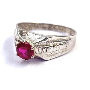 May include: A silver-toned ring featuring a round, faceted, vibrant red gemstone set in a six-prong mounting. The band has a textured, matte finish with a series of vertical rectangular indentations along the side of the shank near the setting.