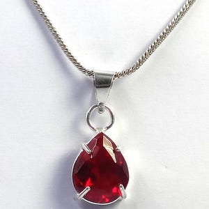 May include: A silver-toned necklace featuring a teardrop-shaped, faceted deep red gemstone pendant. The stone is held in a four-prong setting attached to a simple silver bail, suspended from a delicate, textured silver-toned chain link necklace.