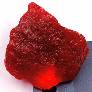 May include: A large, irregularly shaped, translucent red gemstone. The surface has a textured appearance with small, irregular bumps. The gemstone is set against a white background and a dark grey surface.