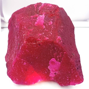 May include: A large, rough, red ruby gemstone. The ruby has a deep, rich red color and a slightly translucent appearance.