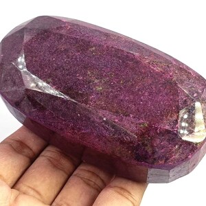 May include: A large, oval-shaped, faceted gemstone with a deep magenta and dark purple colour. The stone features a polished, multi-faceted surface that reflects light, showcasing a textured, crystalline interior with visible natural inclusions.