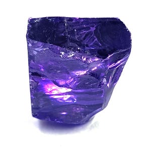 Puede incluir: Una piedra preciosa en bruto de color morado intenso con una estructura cristalina translúcida. La piedra presenta bordes irregulares y dentados, con una superficie lisa y reflectante que muestra fracturas internas visibles.