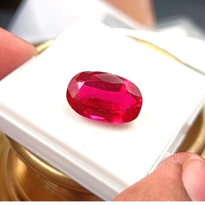 Può includere: Una gemma rossa vibrante, sfaccettata, di forma ovale. La gemma è posta su una superficie bianca, tenuta dalle dita di una persona. Il colore della gemma è un rosso intenso e saturo, con molteplici sfaccettature che catturano la luce.