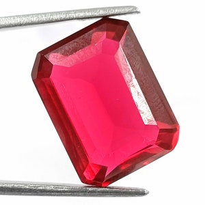 May include: A vibrant red gemstone with a rectangular emerald cut. The gemstone is held by a pair of tweezers.
