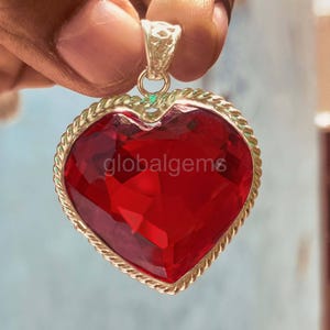 May include: A heart-shaped pendant with a large, faceted red gemstone. The gemstone is set in a gold-colored metal frame with a rope-like border. The text "globalgems" is visible on the gemstone.