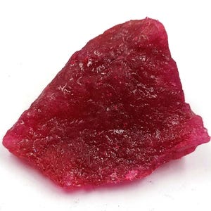 May include: A raw, unpolished gemstone with a deep, vibrant magenta-red color. The stone features an irregular, angular shape with a textured, crystalline surface that catches light, displaying varying levels of translucency throughout.