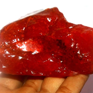 May include: A large, raw, translucent red mineral specimen with an irregular, jagged shape and a glossy, reflective surface. The stone displays deep crimson hues and internal crystalline textures throughout its solid, unpolished structure.