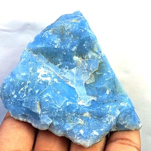 May include: A raw, triangular-shaped piece of blue opal mineral with a translucent, crystalline texture. The stone features a vibrant sky-blue color with visible white inclusions, jagged edges, and a glossy, uneven surface throughout the specimen.
