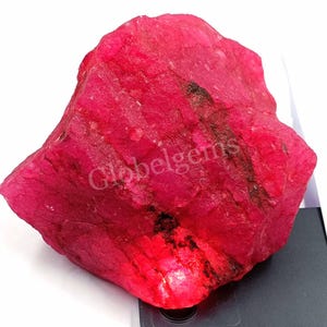 Puede incluir: Una gran gema de rubí rosa en bruto. El rubí tiene un color profundo y rico y es parcialmente translúcido. La piedra está sobre una superficie negra.