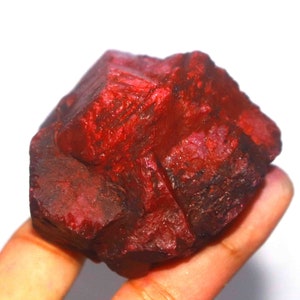 May include: A raw, deep red ruby crystal with a rough, multifaceted surface. The stone displays a rich, saturated crimson hue with natural variations in texture and opacity across its irregular, angular geometric form and crystalline structure.