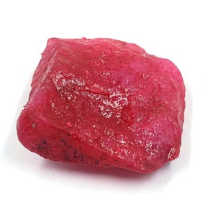 May include: A raw, unpolished gemstone with a vibrant, deep pinkish-red hue. The stone features an irregular, blocky shape with a textured, crystalline surface that displays varying levels of translucency and natural mineral inclusions.