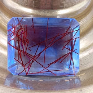 May include: A rectangular, transparent gemstone with a blue hue and red, linear inclusions. The gemstone is faceted with a polished surface, showcasing the internal red lines. The gem is set against a gold-toned background.