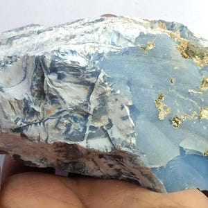May include: A large, rough-hewn stone with a light blue hue, accented by patches of gold and white. The rock's surface has a textured appearance, with visible layers and variations in color. The stone is held in a hand, showcasing its size and natural beauty.