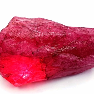 May include: A large, rough, red ruby gemstone. The ruby has a deep, rich red color and is translucent. The gemstone is unpolished and has a natural, irregular shape.