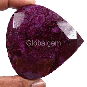 Puede incluir: Una gema facetada en forma de lágrima con un tono magenta intenso. La piedra tiene una apariencia texturizada con vetas más oscuras y el texto "Globalgem" impreso en la superficie. La gema es sostenida por una mano.