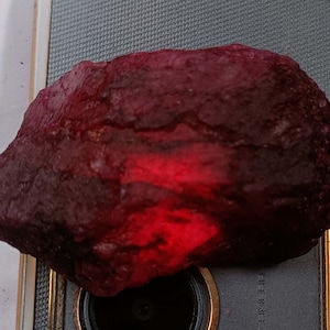 May include: A large, rough, dark red gemstone with a lighter red hue in the center. The gemstone is sitting on a black surface.